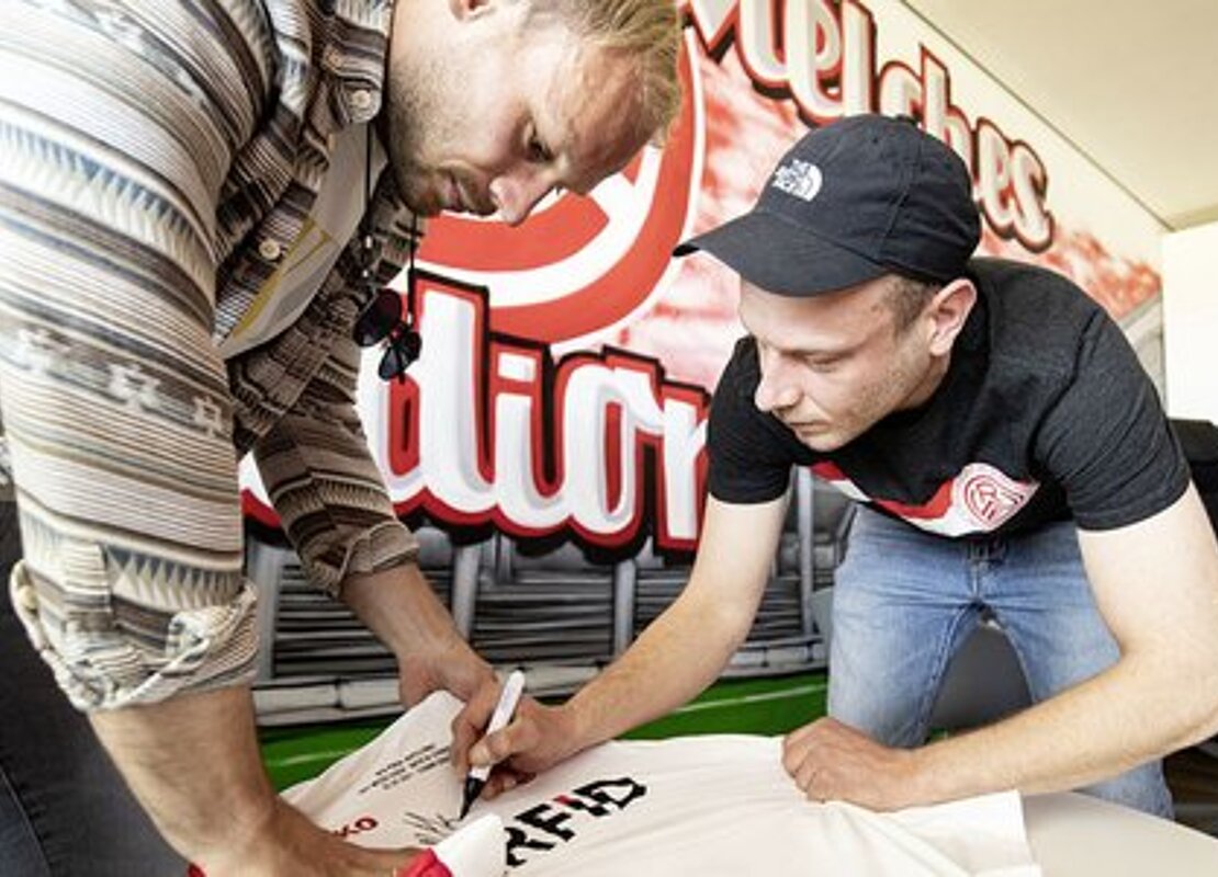 RWE-Linksverteidiger Felix Herzenbruch (links) und Rapper Mike unterzeichnen Trikot der "257ers", das auf Initiative der Allbau GmbH für einen guten Zweck versteigert wird