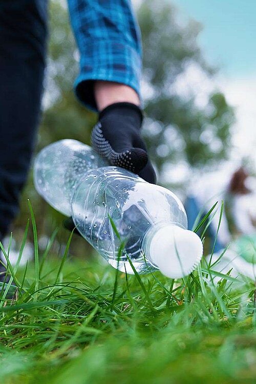 Hand mit Handschuh sammelt leere Plastikflasche vom Rasen auf 