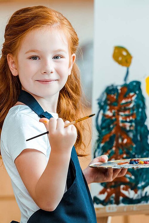 Rothaariges Mädchen malt auf Leinwand, guckt lachend in die Kamera