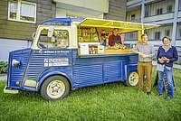 Zwei Personen davor und eine Person in dem geöffneten AllbauCafeMobil, ein restaurierter blauer Citroen-Marktwagen, aus dem Getränke und Snacks verkauft werden