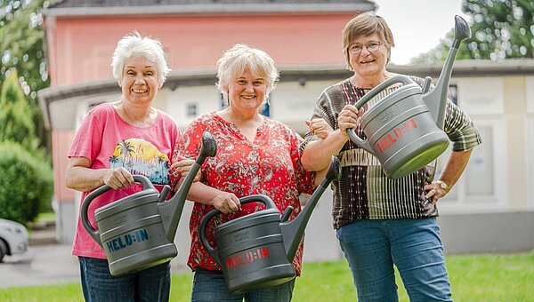 Drei Seniorinnen mit Gießkannen auf einer Wiese vor einem Haus