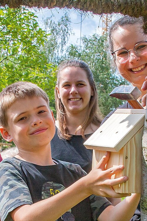 Die Kinderbeauftragte Anna Golz der Allbau GmbH (Mitte) schaut zu, wie zwei Kinder ein Vogelhäuschen aufhängen